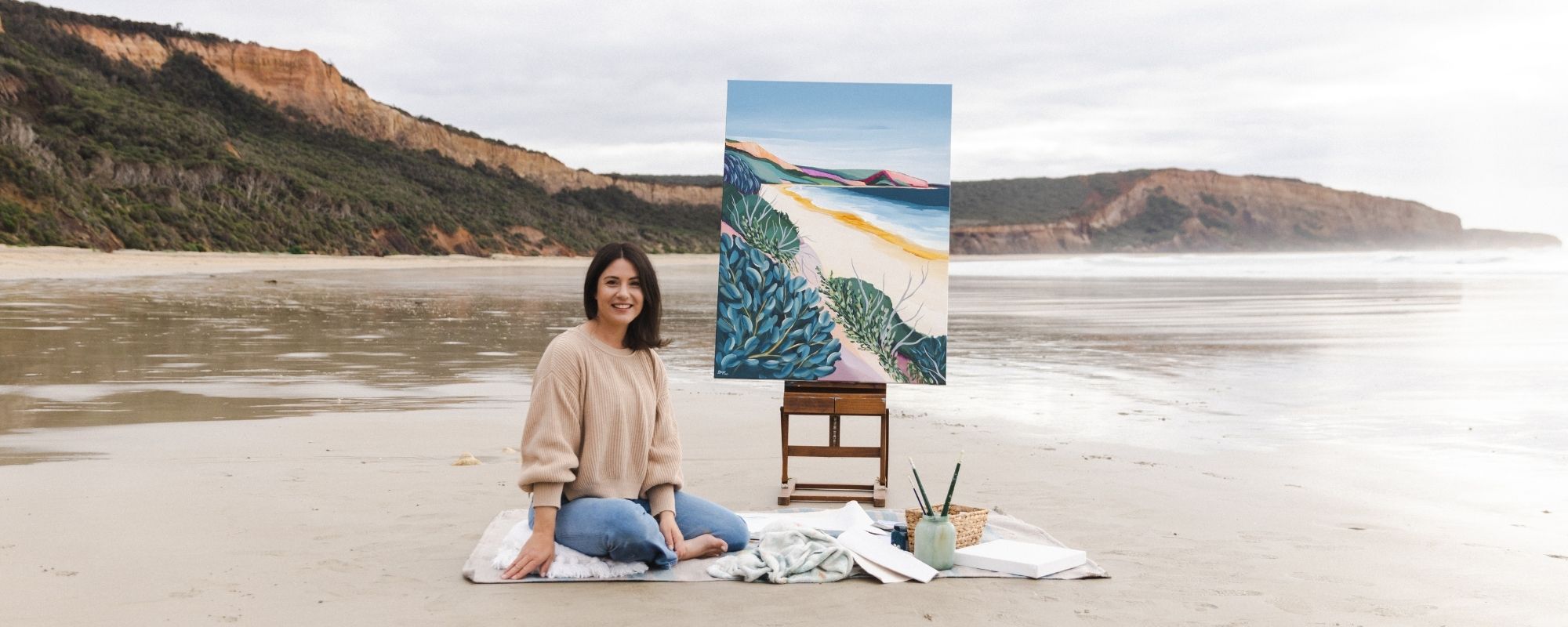 Australian artist Lauren Danger sitting on a blanket with a painting of a beach scene on a stand, surrounded the location Point Addis on the Great Ocean Road, Surf Coast, Victoria, Australia