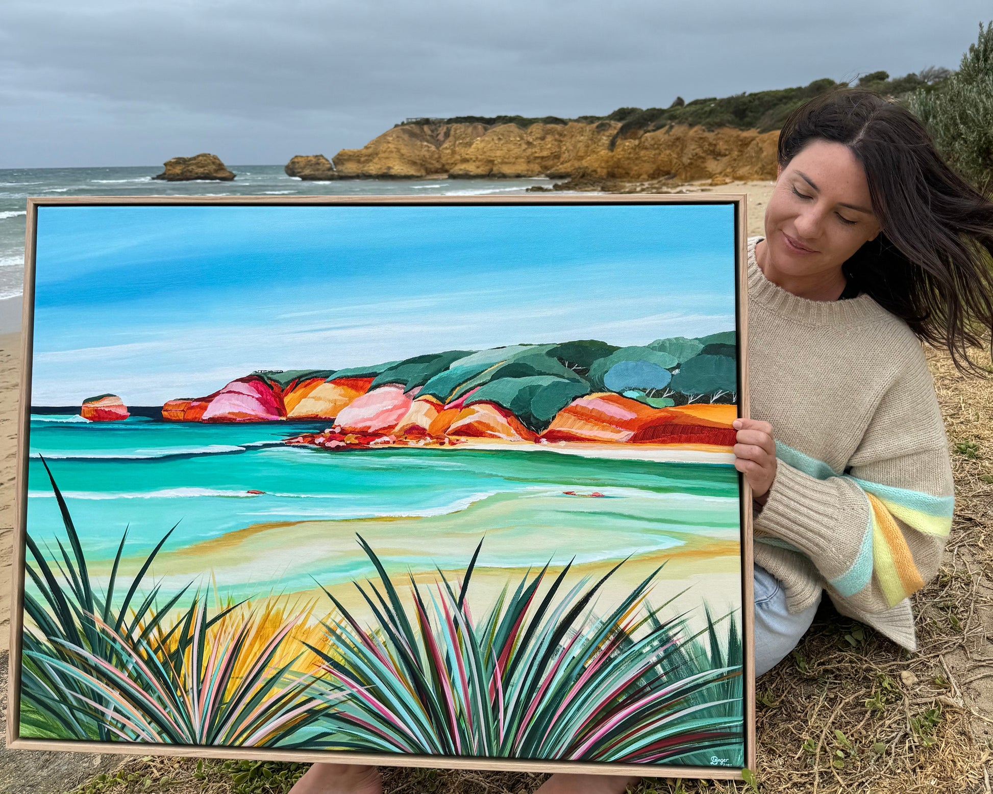 Australian artist Lauren Danger holding a colourful coastal artwork of raw oak framed canvas limited edition print of Rocky Point no. 3.  Photo taken on location, Torquay, Surf Coast, Great Ocean Road Australia.