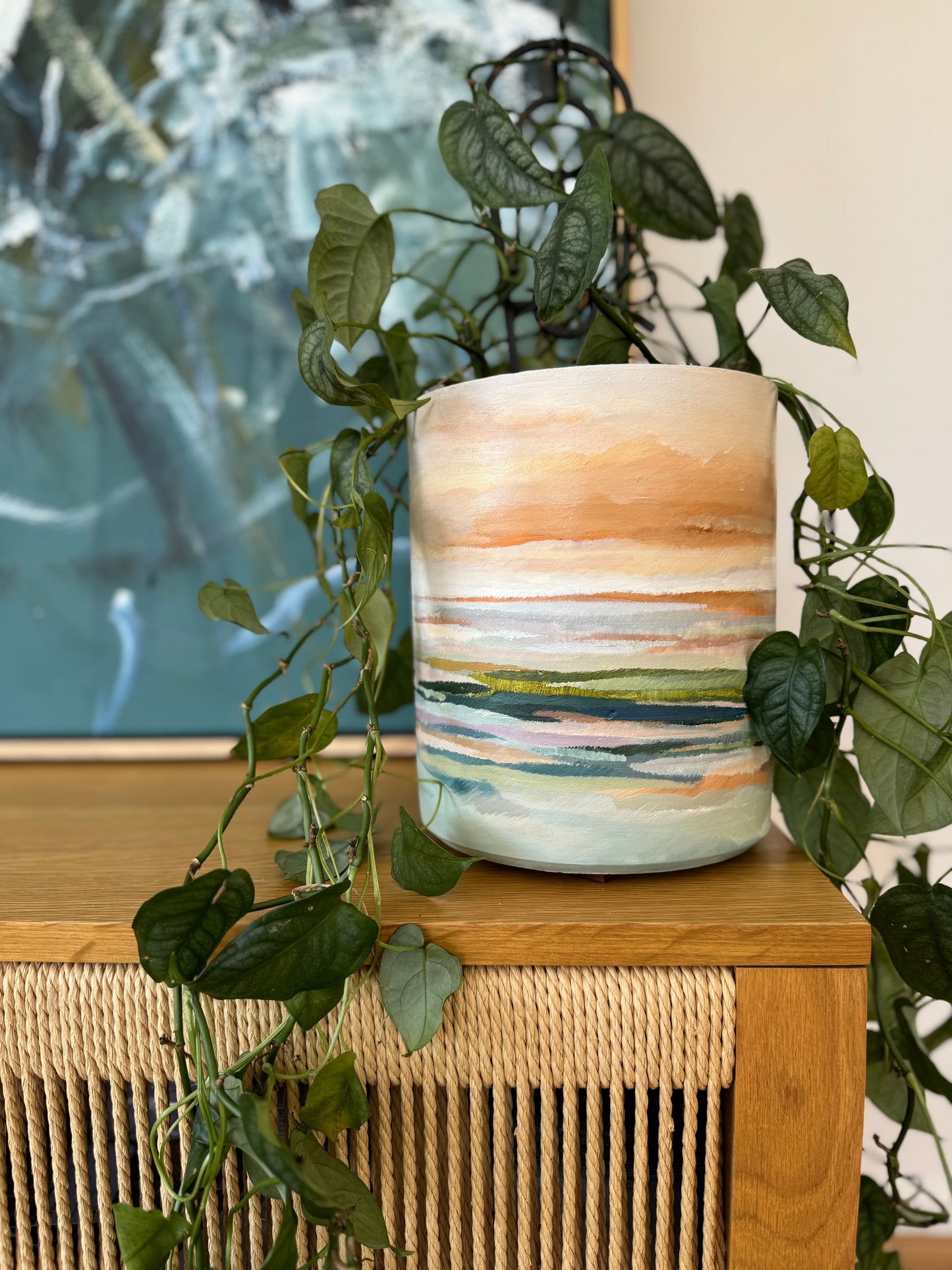 Hand-painted ceramic vase with pastel coastal landscape, displayed on timber sideboard with trailing greenery